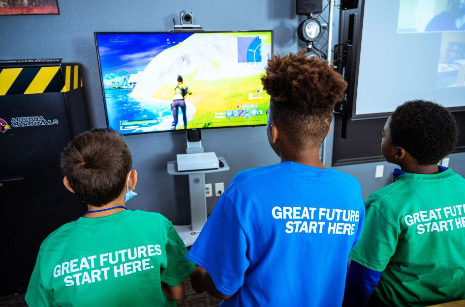 Three Boys & Girls Club members play a video game together, wearing shirts with 'Great Futures Start Here' printed on the back.